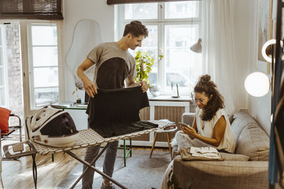 Man ironing clothes looking at woman using smart phone on sofa at home