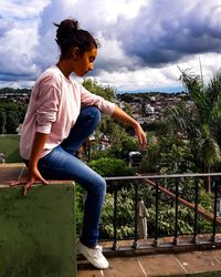 Thoughtful teenage girl sitting on terrace against cloudy sky