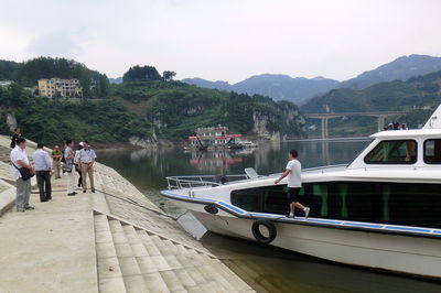 People looking at yacht moored at riverbank by mountains against clear sky