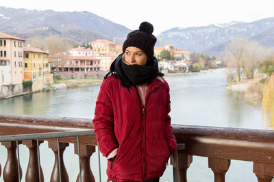Portrait of young woman standing on railing against river during winter