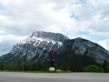 Happy woman with arms raised standing at roadside against snowcapped mountains