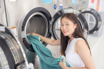 In the self-service laundry with dryer machines in the backdrop, a young woman enjoys clean ironed.