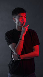 Young man with finger on lips looking away while standing against black background