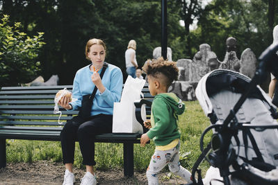 Mother sitting on bench and having food while looking at son running in park
