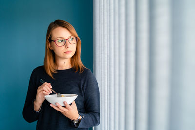Young woman holding food in bowl while standing against wall