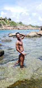 Portrait of shirtless young man standing in sea against cloudy sky