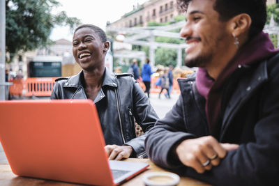 Young woman with laptop smiling while sitting by friend at sidewalk cafe