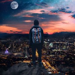 Rear view of man standing by illuminated buildings against sky at sunset