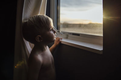 Thoughtful shirtless boy looking through window at home during sunset