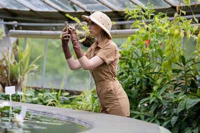 Side view of woman photographing while standing by plants 