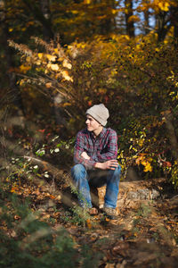 Teen boy sits alone on log in woods and smiles with arms crossed
