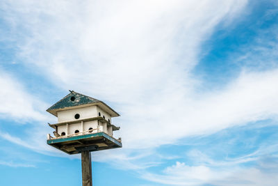 Low angle view of  large birdhouse in need of repair, a bird perched set against a whispy sky. 