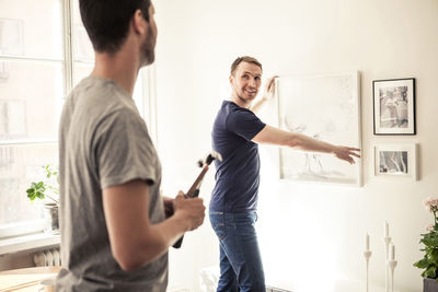 Side view of young gay man looking at partner while hanging frame on wall in home