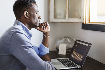 Thoughtful man looking away while sitting with laptop computer at home