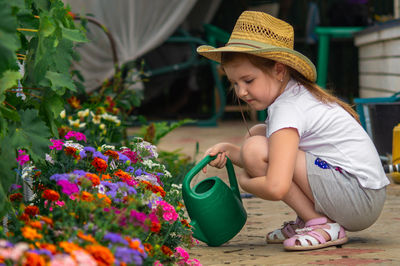 Lovely little girl outdoor in garden takes care of flower bed by watering it by green watering can. 