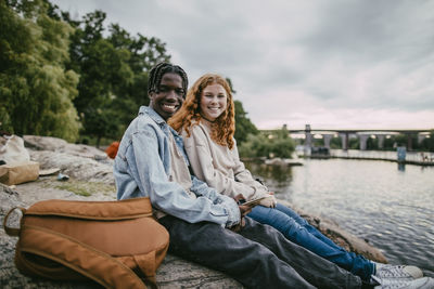 Portrait of smiling multiracial male and female friend sitting on rock near sea