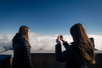 Woman photographing friend at observation point against blue sky