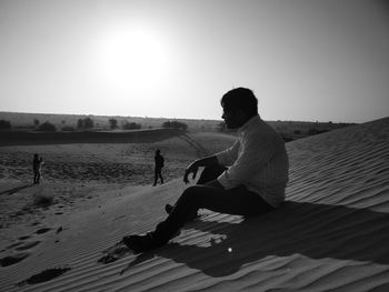 Man sitting on sand at desert against sky