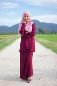 Portrait of young woman in hijab walking on footpath against sky