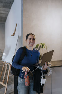Portrait of happy businesswoman with laptop and mobile phone charger standing at workplace