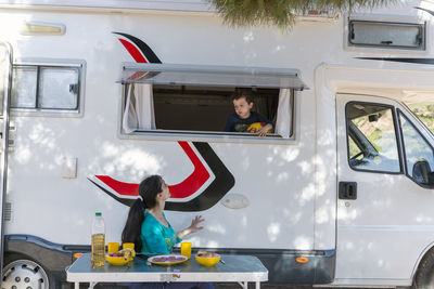 Woman at camping table next to her motor-home and child in the window