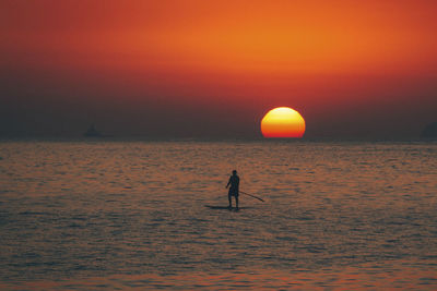 Silhouette man rowing boat on sea against orange sky