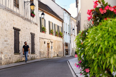 A man in a casual outfit walks down a street with stone houses and flowers in the windows.