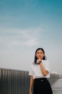 Portrait of smiling young woman gesturing thumbs up while standing by railing against sky