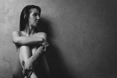 Naked woman sitting by wall while looking away