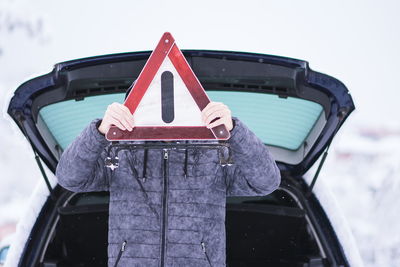 Man wearing warm clothing holding warning sign against car during winter