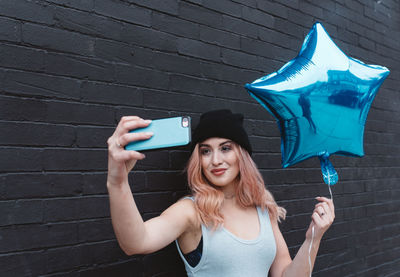 Smiling woman taking selfie through smart phone while holding blue star shape balloon by wall