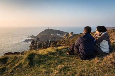 Rear view of couple sitting on rock by sea against clear sky