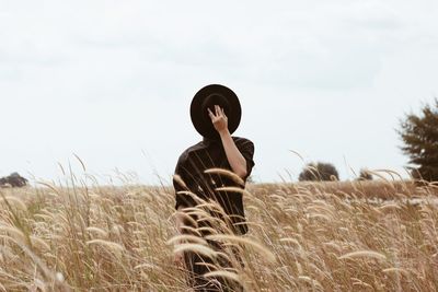 Man hiding face with hat while standing on field against clear sky