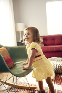 Curious girl looking away while standing by chair at home