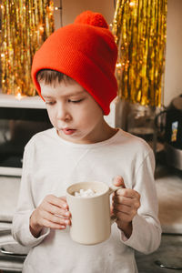 Boy in a white t-shirt and a red hat drinking cocoa with marshmallows in the kitchen.