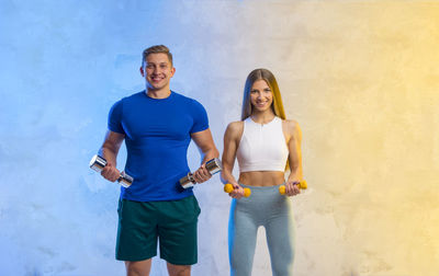 Portrait of young couple holding dumbbell standing against illuminated wall