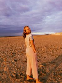 Full length rear view portrait of young woman standing on beach against cloudy sky