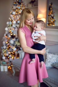 Woman in a pink dress holds her son's child in her arms standing at the christmas tree in the studio