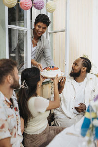 Smiling young man giving cake to male and female friends sitting at cafe