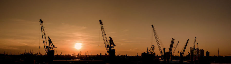 Silhouette cranes at harbor against sky during sunset