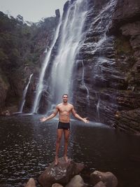 Full length of shirtless man standing against waterfall