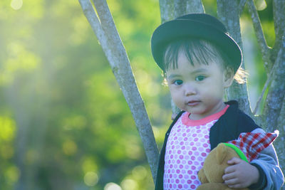 Portrait of cute girl holding teddy bear while standing against tree