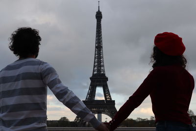 Rear view of couple holding hands while looking at eiffel tower against cloudy sky