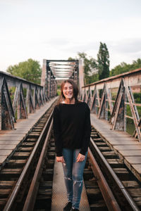 Portrait of young woman standing on footbridge against sky