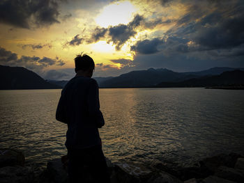 Rear view of man standing at beach against sky during sunset