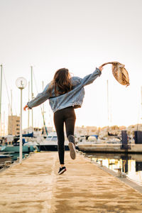 Rear view of young woman jumping at harbor during sunset