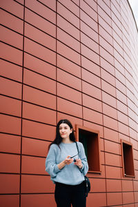 Young woman using phone while standing against wall
