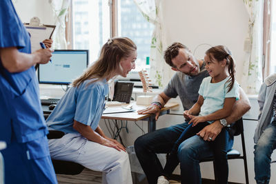 Female pediatrician talking to smiling girl sitting with father in medical examination room