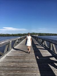 Rear view full length of woman walking on boardwalk over lake against blue sky