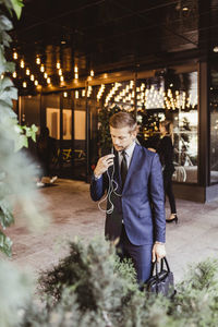 Male entrepreneur using in-ear headphones while standing by plants